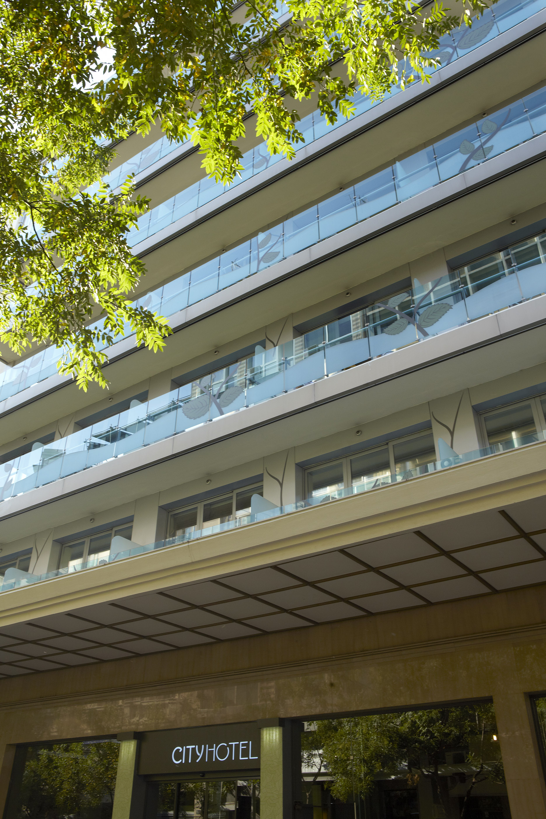 
City Hotel exterior view framed by leafy trees, showcasing modern glass balconies and the main entrance with illuminated signage