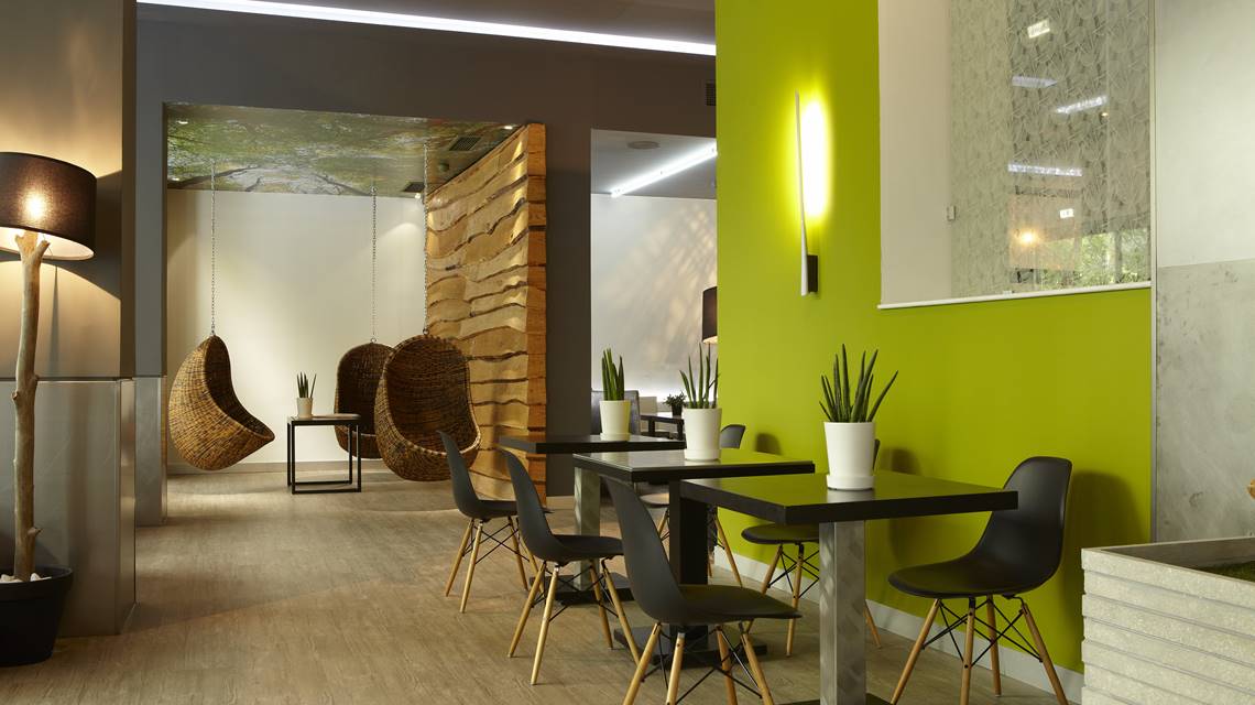 City Hotel indoor public space with modern black tables, minimalist chairs, potted plants, and inviting wicker hanging chairs, all set against a bright green accent wall and natural wood decor