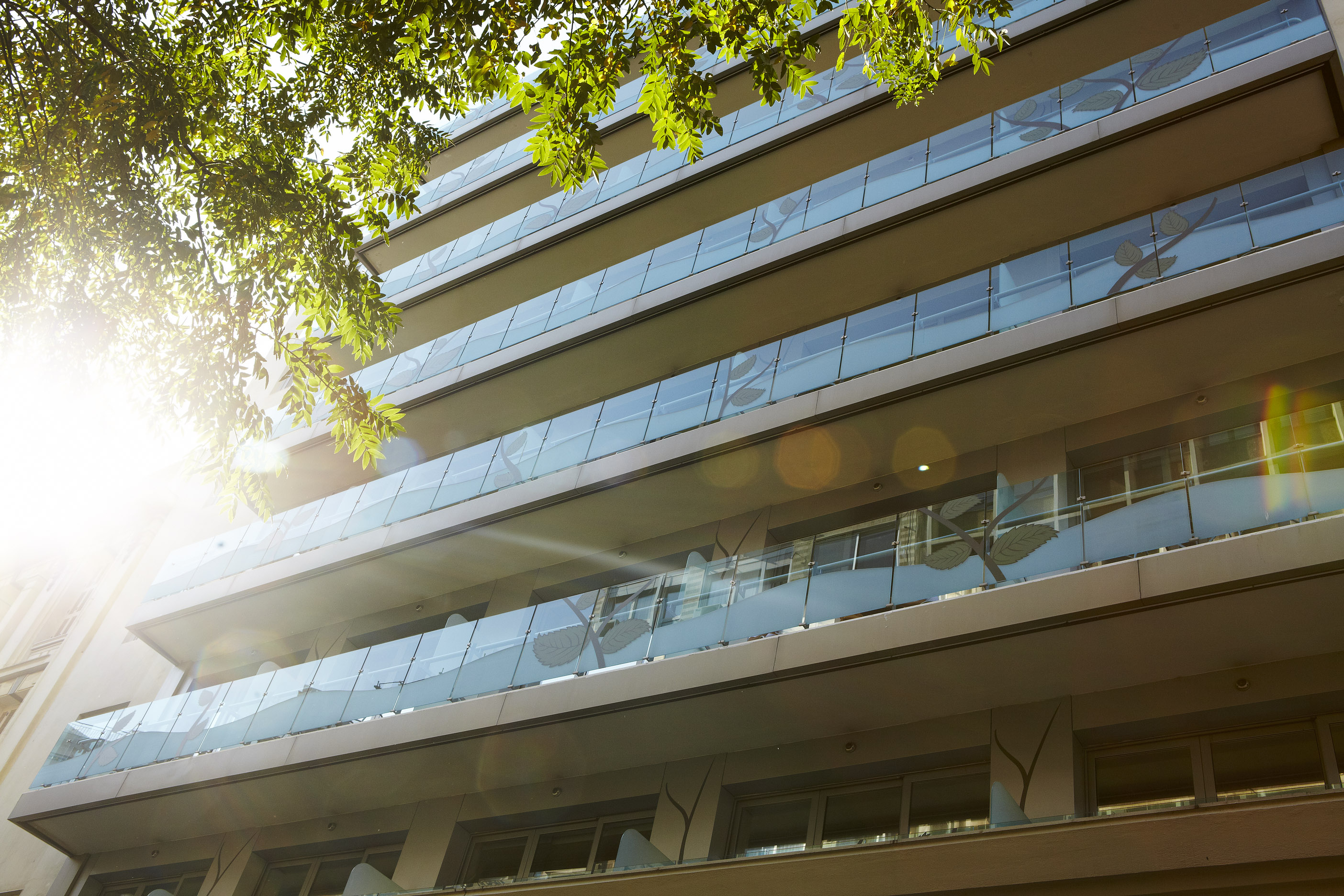 
City Hotel exterior view from ground level, highlighting sunlit balconies with decorative glass panels and leafy urban surroundings
