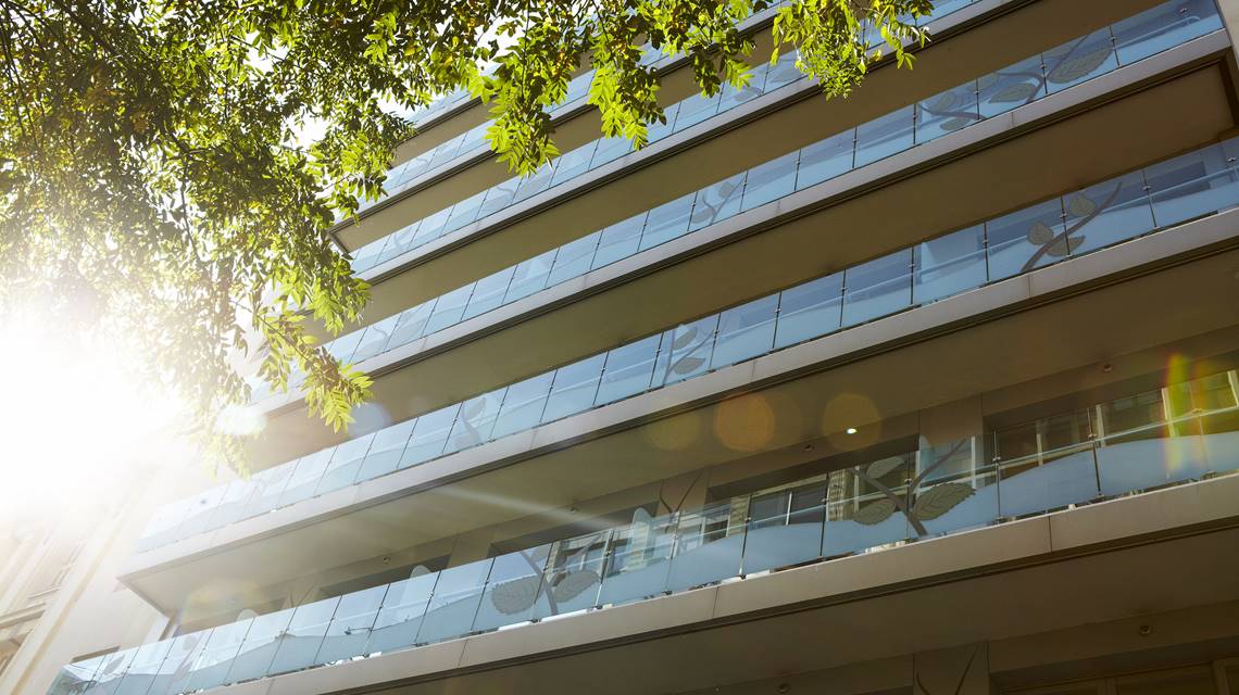 City Hotel exterior view from ground level, highlighting sunlit balconies with decorative glass panels and leafy urban surroundings
